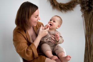 Mother smilingly holds a baby on her lap, feeding or playing with the baby's fingers indoors.